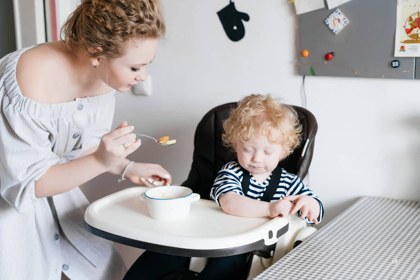 curly young woman mother feeds her son with useful food, family dinner in the kitchen - Photo, image