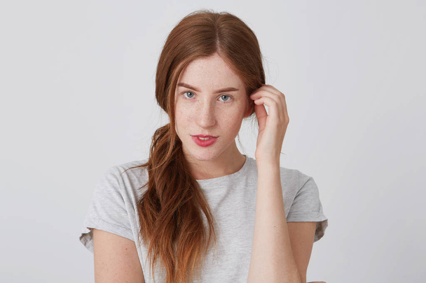 Портрет красивой молодой женщины с длинными рыжими волосами и веснушками в футболке, позирующей и смотрящей на камеру на белом фоне
 - Фото, изображение