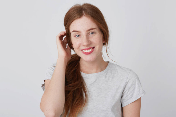 Mladá žena šťastná krásná zrzka s pihy dotýká jejích vlasů a usmívá izolovaná bílém zeď Looking na kameru - Fotografie, Obrázek