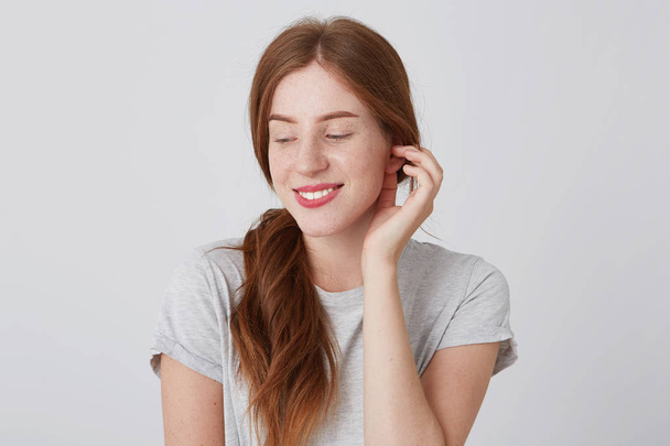 Closeup of happy playful young woman with long red hair and freckles touching her ear and smiling isolated over white background Looking to the side - Fotó, kép