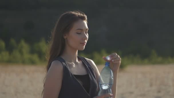 Jonge vrouw reiziger drinkwater uit een plastic fles in de natuur op hete zanderig veld. Meisje gevoel dorst met water onderbreking bij zonsondergang - Video