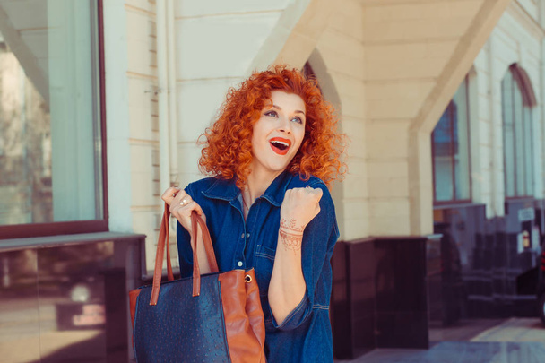 Happiness, winner. Portrait smiling beautiful city lifestyle woman looking up raising hand pumping fist. Pretty young girl with red curly hair isolated street background. Positive expression - Foto, Bild