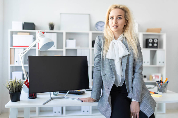 Young girl stands in office, leaning on desk. Nearby is a monitor. - Foto, Bild