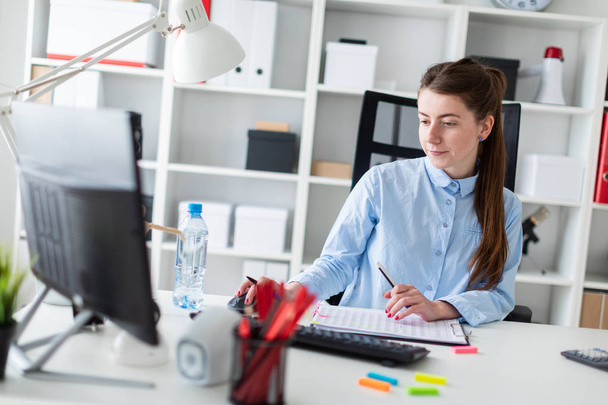 Молода дівчина сидить за столом в офісі, тримає ручку в руці і працює за комп'ютером
. - Фото, зображення