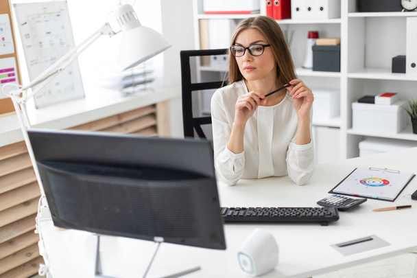 Una joven está sentada en el escritorio de la oficina, sosteniendo un lápiz en la mano y mirando al monitor
. - Foto, imagen