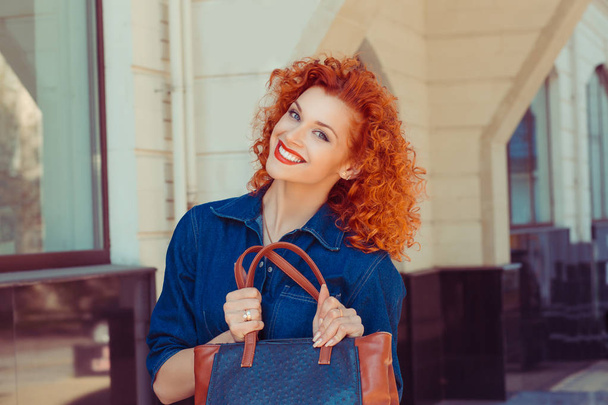 Happy handsome beautiful woman holding showing her new orange leather jeans bag, looking camera. Pretty young girl with red curly hair, isolated street background. Positive facial expression - Zdjęcie, obraz