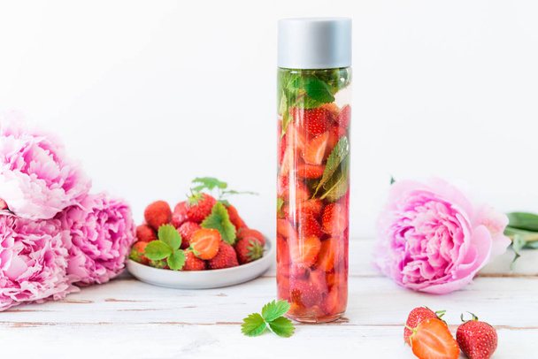 Eau infusée de désintoxication aux fraises et menthe dans une bouteille de sport avec des fraises et des pivoines à proximité sur fond clair
 - Photo, image