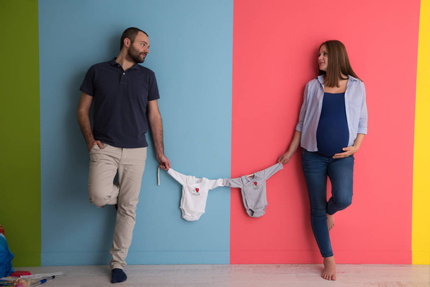 Mooie zwangere vrouw en haar echtgenoot verwachten baby baby bodysuits houden en lachend over kleurrijke achtergrond - Foto, afbeelding
