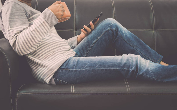 Жінка на шкіряному дивані використовує мобільний телефон і тримає чашку кави вдома
. - Фото, зображення
