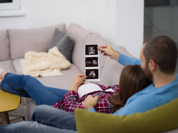 Young pregnant couple looking baby's ultrasound photo while relaxing on sofa at home - Foto, Imagen