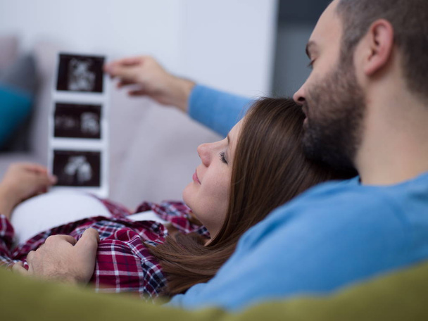 Young pregnant couple looking baby's ultrasound photo while relaxing on sofa at home - Fotografie, Obrázek