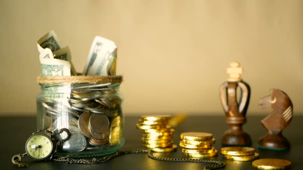 Εξοικονόμηση χρημάτων κέρματος σε βάζο. Σύμβολο της επένδυσης, κρατώντας τα χρήματα ιδέα. Συλλογή μετρητών τραπεζογραμμάτια σε γυαλί κασσίτερο. Αμερικανικά δολάρια με αυξανόμενη στήλες των χρυσών νομισμάτων σε μαύρο με ρετρό ρολόι, σκάκι. - Πλάνα, βίντεο