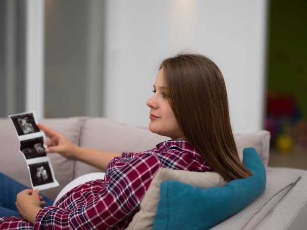 Young pregnant woman looking baby's ultrasound photo while relaxing on sofa at home - Φωτογραφία, εικόνα