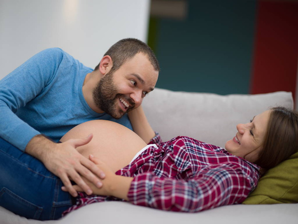 Happy future dad listening the belly of his pregnant wife while relaxing on sofa at home - Фото, зображення