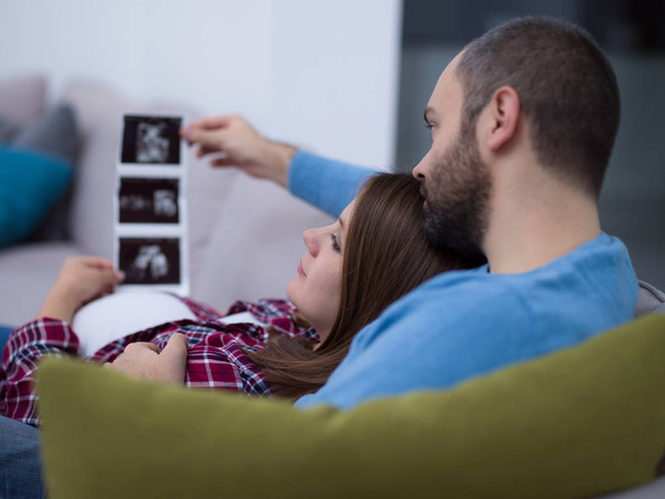 Young pregnant couple looking baby's ultrasound photo while relaxing on sofa at home - Фото, зображення