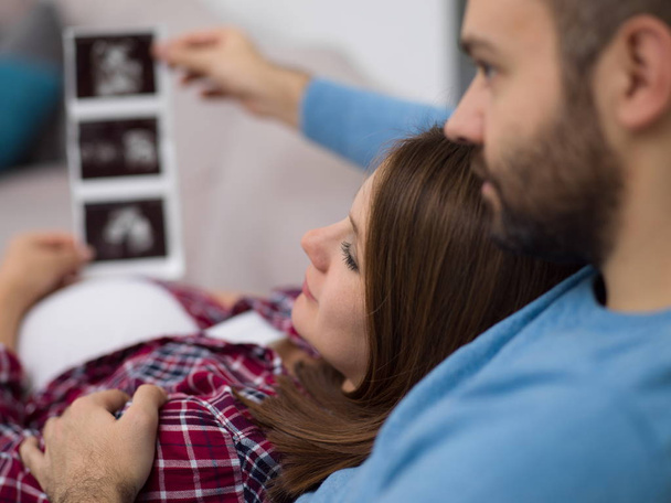Young pregnant couple looking baby's ultrasound photo while relaxing on sofa at home - Foto, afbeelding