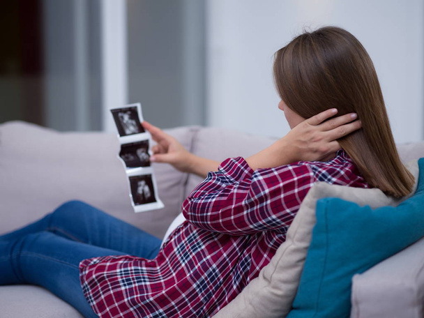 Young pregnant woman looking baby's ultrasound photo while relaxing on sofa at home - Valokuva, kuva