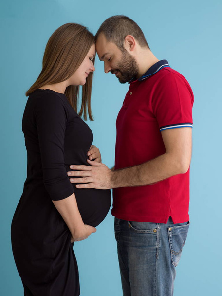 Portrait of a happy young couple,man holding his pregnant wife belly isolated over blue background - 写真・画像