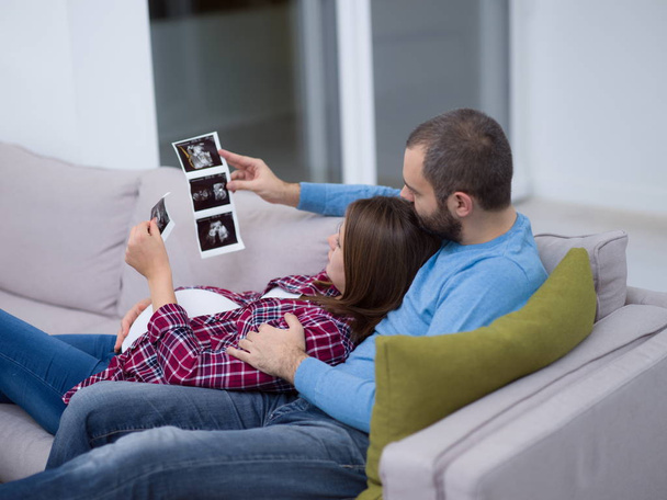 Young pregnant couple looking baby's ultrasound photo while relaxing on sofa at home - Foto, imagen