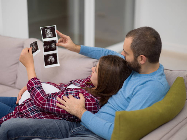 Young pregnant couple looking baby's ultrasound photo while relaxing on sofa at home - Φωτογραφία, εικόνα