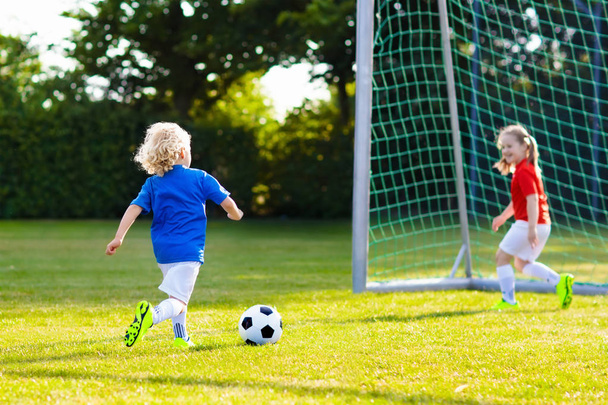 子供たちは野外でサッカーをする。子供たちはサッカーの試合でゴールを決めた。女の子と男の子はボールを蹴る。チームジャージとクリートで子供を走らせている。サッカー部所属。若い選手のためのスポーツトレーニング. - 写真・画像