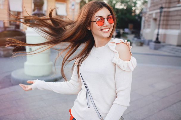 Close up image of happy brunette woman in sunglasses and autumn clothes posing sideways outdoors.Happy woman in sunglasses and dress walking outdoors. Looking at camera. the wind fluttering hair. - Фото, изображение