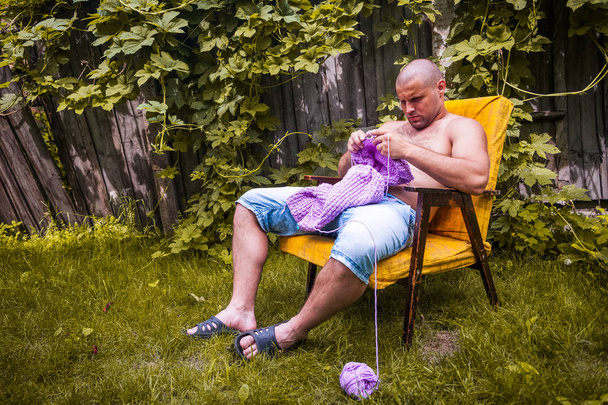 Mladý holohlavý muž sedí na křesle a plete svetr z přírodních lila vln ěných vláken na chatě, v pozadí dřevěný plot, hroznové keře - Fotografie, Obrázek
