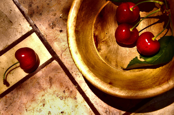 Zenith view of still life of three cherries into wood bowl and a lonely one at the other side, all over stone table with lines crossing - Photo, Image