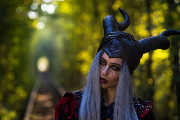 terrible woman with horns and amazing makeup in the tunnel of forest with rails close up - Photo, image