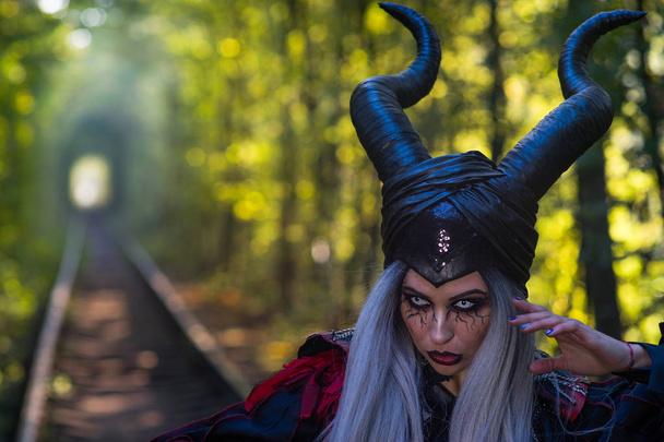 terrible woman with horns and amazing makeup in the tunnel of forest with rails close up - Fotó, kép