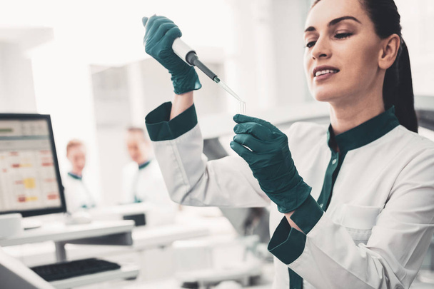 Investigation. Portrait of positive concentrated laboratory assistant using pipette and taking analyses while holding glass tube - Foto, imagen