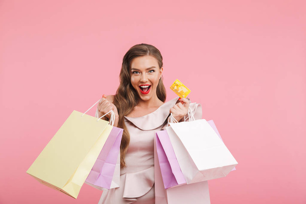 Foto bohatých módní šaty usmívající se žena 20s zatímco drží různé nákupní tašky a kreditní karty v rukou izolované růžové pozadí - Fotografie, Obrázek