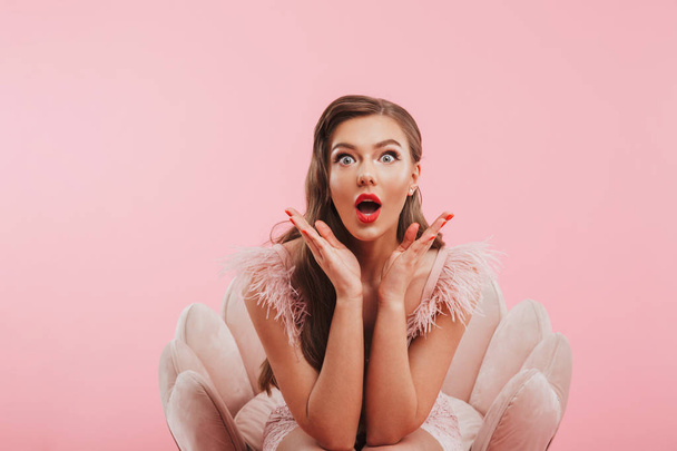 Portrait of beautiful excited woman in dress expressing surprise while sitting in armchair isolated over pink background - Foto, immagini