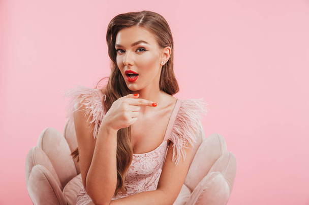 Portrait closeup of attractive young woman in dress sitting in armchair and pointing finger aside on copyspace isolated over pink background - Foto, immagini