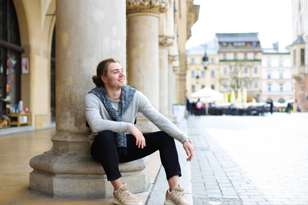 Młody kaukaski mężczyzna turystycznych siedzi w pobliżu kolumn na rynku głównym w Krakowie. - Zdjęcie, obraz