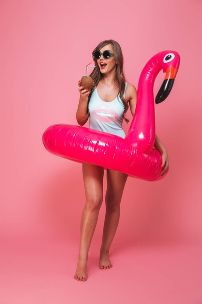Full length portrait of a pretty young woman dressed in swimsuit posing with inflatable flamingo and coconut cocktail isolated over pink background - Foto, Imagem
