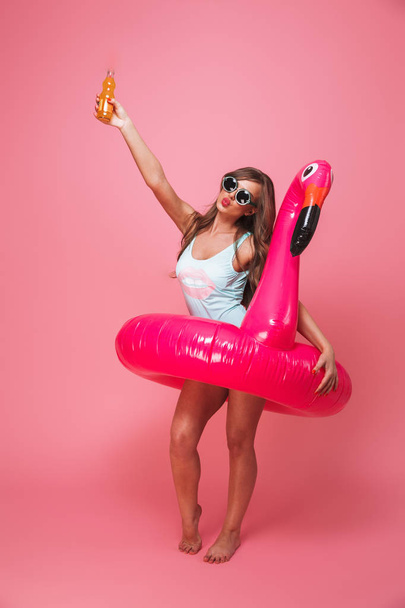 Full length portrait of a happy young woman dressed in swimsuit posing with inflatable flamingo and soda bottle isolated over pink background - Foto, afbeelding