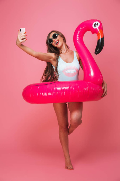 Full length portrait of a pretty young woman dressed in swimsuit posing with inflatable flamingo while taking a selfie isolated over pink background - Foto, afbeelding