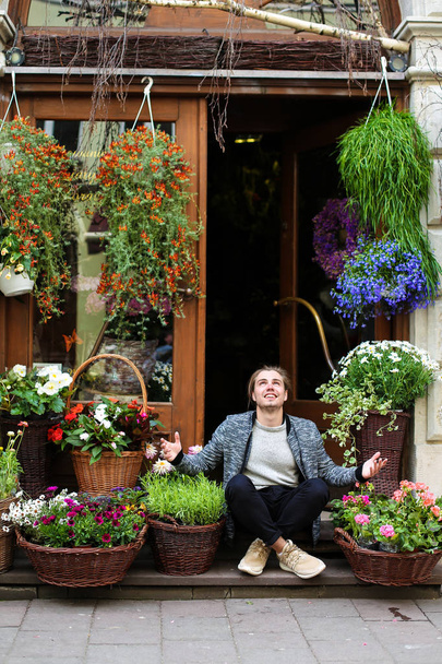 Joven hombre caucásico sentado en la floristería cerca de ramos en cestas
. - Foto, Imagen