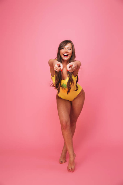 Full length portrait of a smiling young woman dressed in swimsuit pointing fingers at camera isolated over pink background - Foto, afbeelding