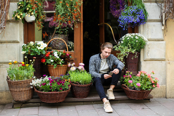 Joven hombre europeo charlando por teléfono inteligente y sentado en la floristería cerca de ramos de flores en cestas
. - Foto, imagen