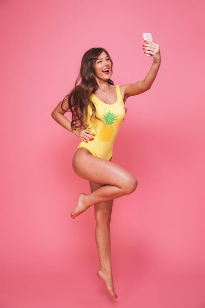 Full length portrait of a joyful young woman dressed in swimsuit taking selfie with mobile phone while jumping isolated over pink background - Photo, Image