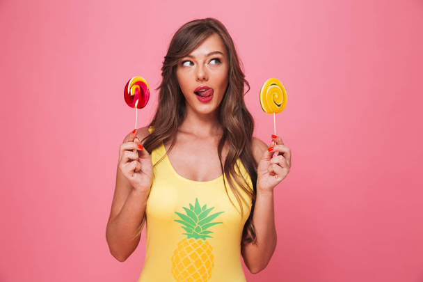 Portrait of a cute young woman dressed in swimsuit holding colorful lollipops isolated over pink background - Foto, immagini