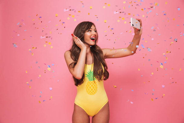 Portrait of a happy young woman dressed in swimsuit taking a selfie under confetti rain isolated over pink background - Photo, Image