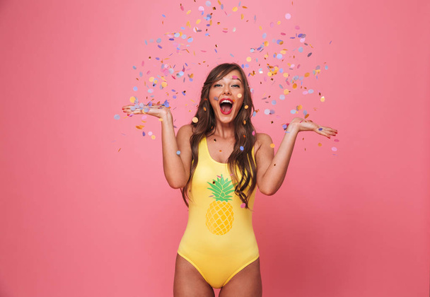 Portrait of a happy young woman dressed in swimsuit having fun under confetti rain isolated over pink background - Foto, Imagen