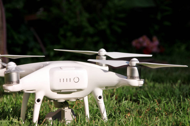 weiße Quadrocopter-Drohne mit 4k Digitalkamera auf Gras ist startbereit, um in der Luft zu fliegen, um Fotos zu machen, Aufnahmen von oben zu machen. Drohne mit vier Motoren, Propellern, Kamera und rotem Warnlicht. - Foto, Bild