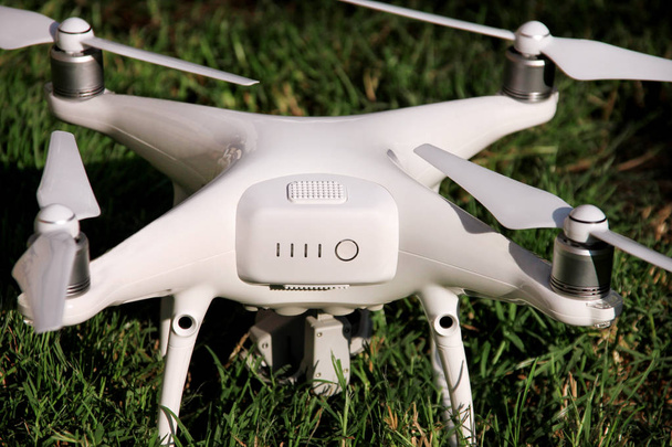 Λευκό τετράγωνο ελικόπτερο Drone με 4 k ψηφιακή φωτογραφική μηχανή στο γρασίδι είναι έτοιμο για απογείωση να πετάξει στον αέρα για να τραβήξετε φωτογραφίες, να καταγράψει στιγμιότυπα από ψηλά. Drone με τέσσερις κινητήρες, έλικες, κάμερα και κόκκινα φώτα προειδοποίησης. - Φωτογραφία, εικόνα