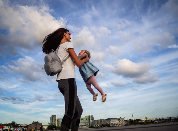 Family values: Mom with sunglasses whirl with a little daughter in a blue dress against the sky and clouds. Authentic family. - 写真・画像