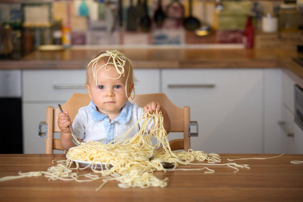 Niño pequeño, niño pequeño, comiendo espaguetis para el almuerzo y haciendo un desastre en casa en la cocina
 - Foto, imagen