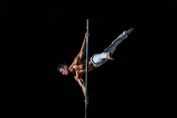 A strong muscular guy dressed in white pants performs a trick on a pole in on a black background - Фото, изображение